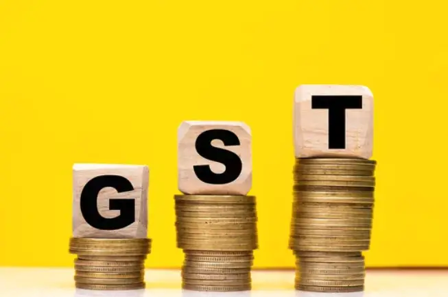 Three wood cubes, each with a letter that spells GST. Each cube is sitting on a pile of coins.